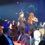 Two violinists playing on a stage surrounded by seated people